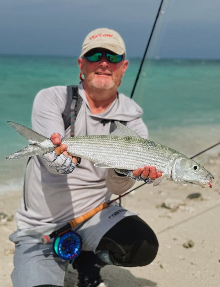 Sportfiskare med solglasögon och kepsar håller stor silverfisk på sandstrand, turkost hav och fiskeutrustning i bakgrunden.