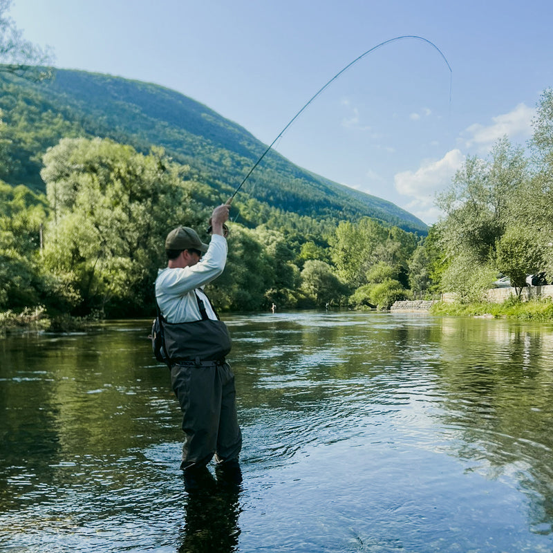 Sportfiskare på fiskeresa i Bosnien vadar i klar älv, kastar lina omgiven av grönska och kullar.